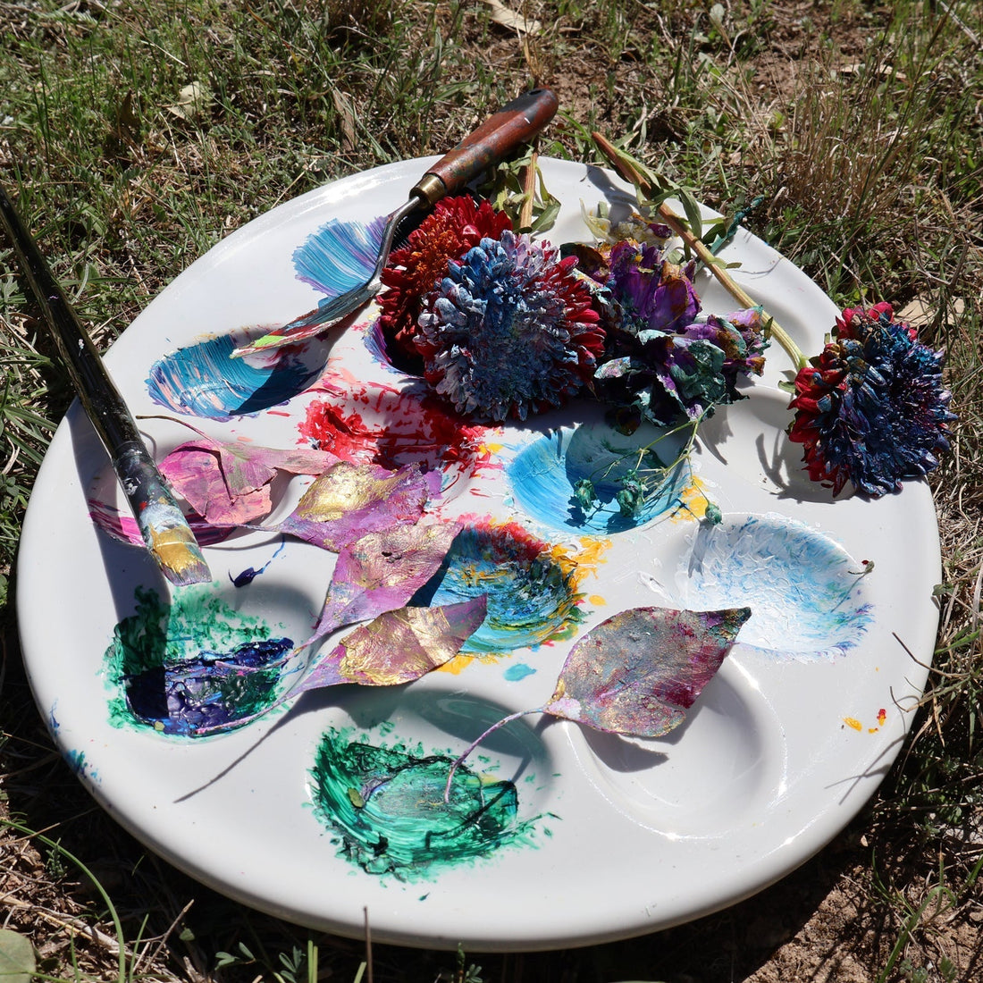 Ceramic egg paint holder with dried paint, painted leaves, painted flowers, and painted art brushes resting on open grass outdoors.