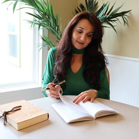 Stephanie Christley, Artist & Founder of SLChristley Designs, sitting and smiling while flipping through her journal in a light-filled room, wearing a long sleeve green blouse with a large window and indoor green tree in the background.