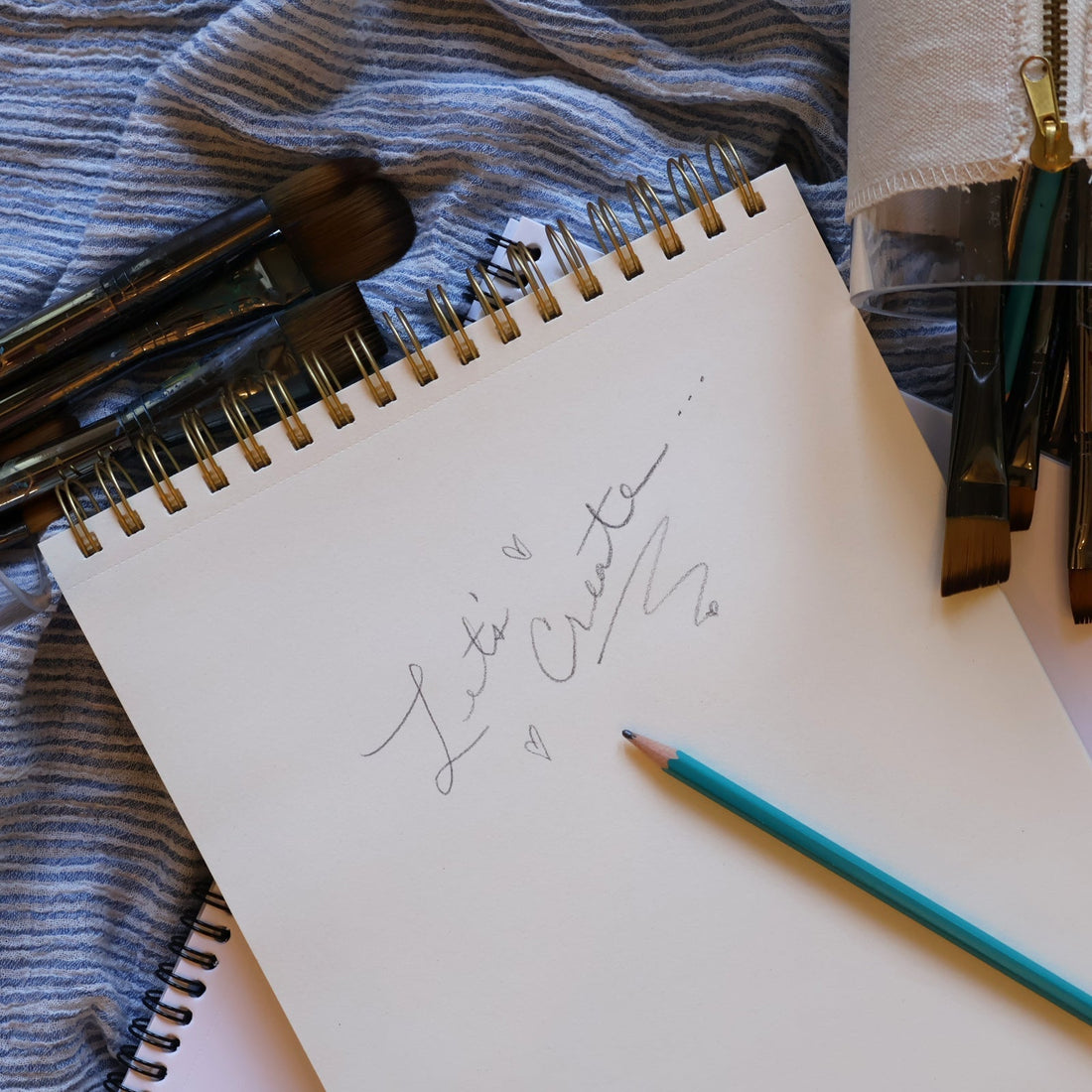 Art supplies, pencils, and brushes laid over a sketchbook with the handwritten phrase, "Let's Create."