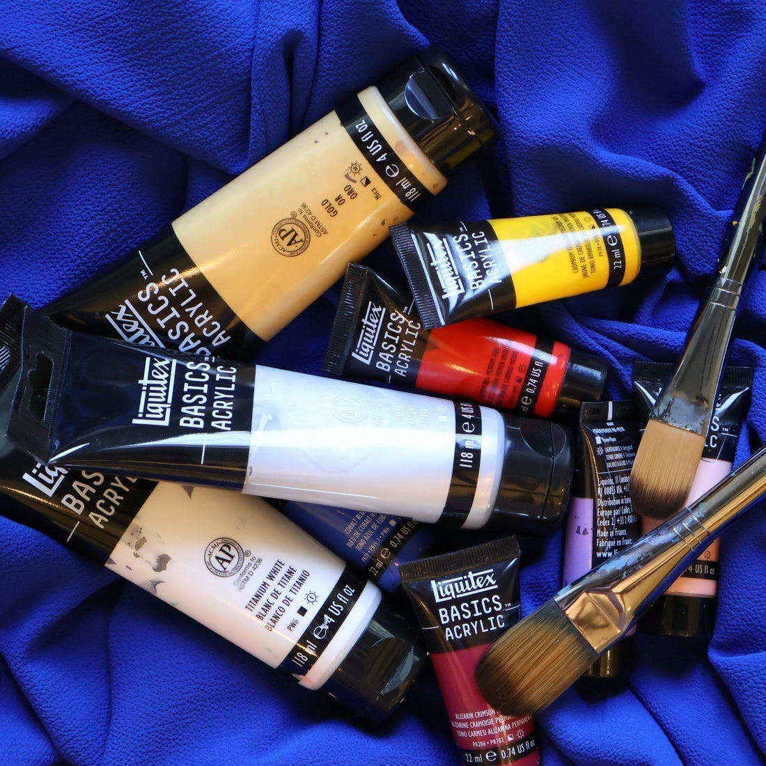Tubes of acrylic paint (silver, white, gold, red, yellow) and paintbrushes piled together on blue fabric.
