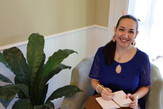 Stephanie Christley, Artist & Founder of SLChristley Designs, sitting indoors smiling and journaling beside a green plant.