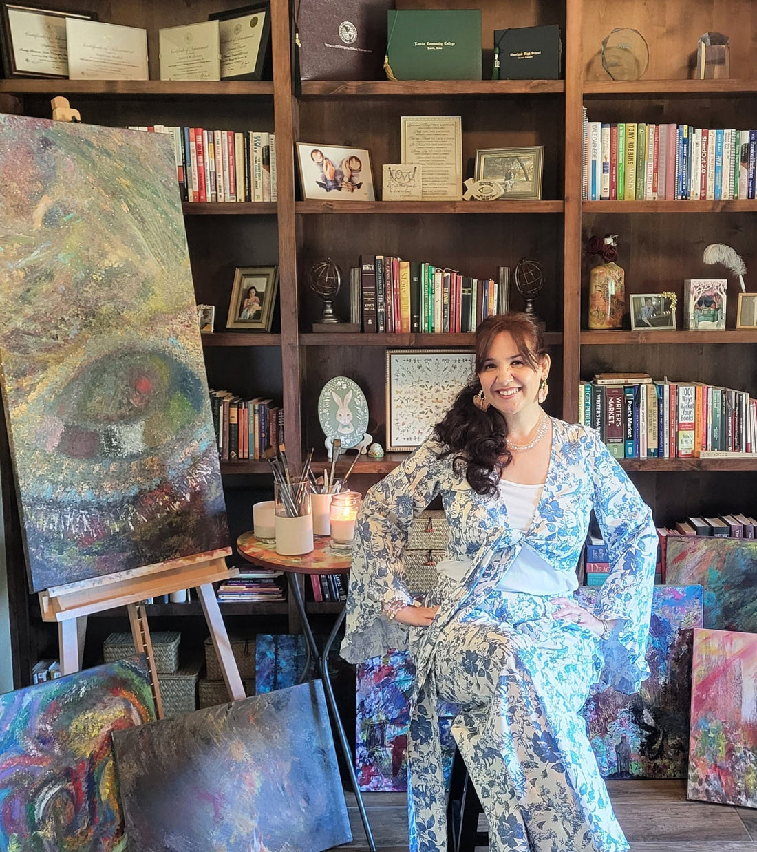 Stephanie Christley, Artist & Founder of SLChristley Designs, sitting on a stool in her study smiling with hands on hips, wearing a blue and white outfit, surrounded by her products, easels, paintings, and a large bookshelf.