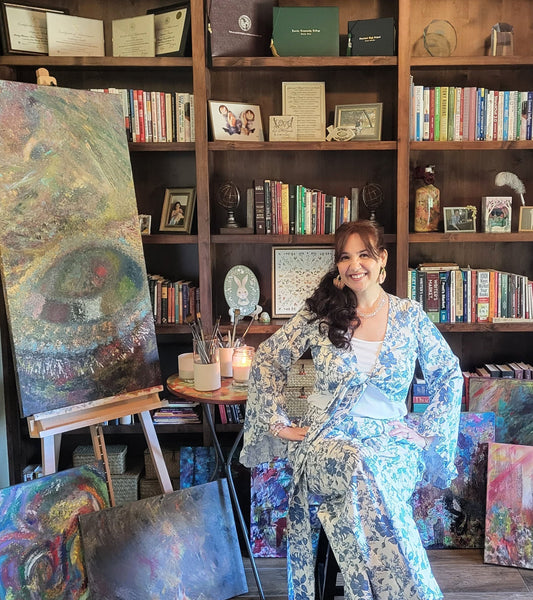 Stephanie Christley, Artist & Founder of SLChristley Designs, sitting on a stool in her study smiling with hands on hips, wearing a blue and white outfit, surrounded by her products, easels, paintings, and a large bookshelf.