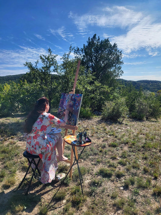 Stephanie Christley, Artist & Founder of SLChristley Designs, painting outdoors in the Texas Hill Country at a tall easel surrounded by green trees, blue sky, and acrylic paints.