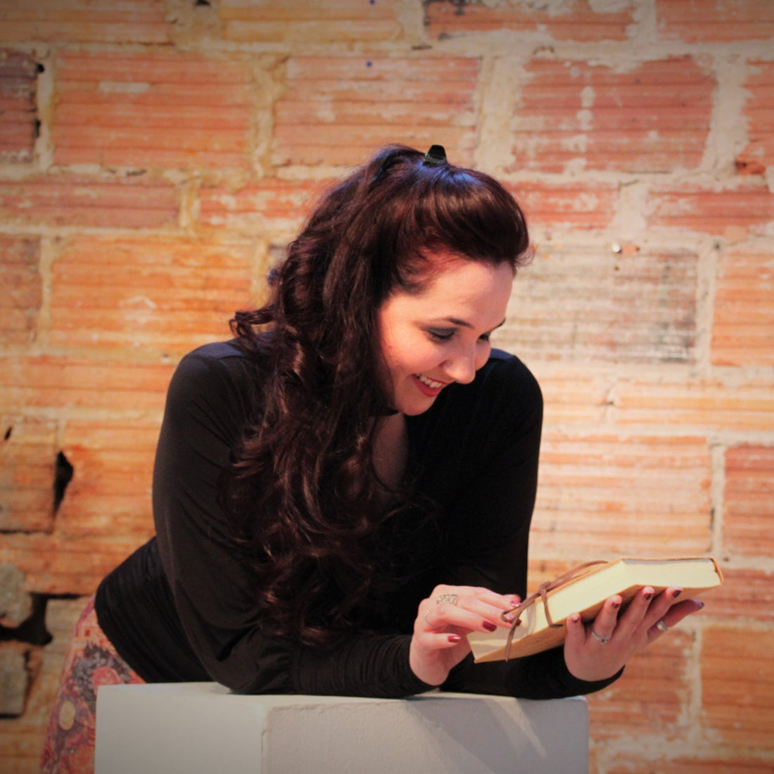 Stephanie Christley, Artist & Founder of SLChristley Designs, smiling and looking down at her journal in front of a lofty orange brick wall.