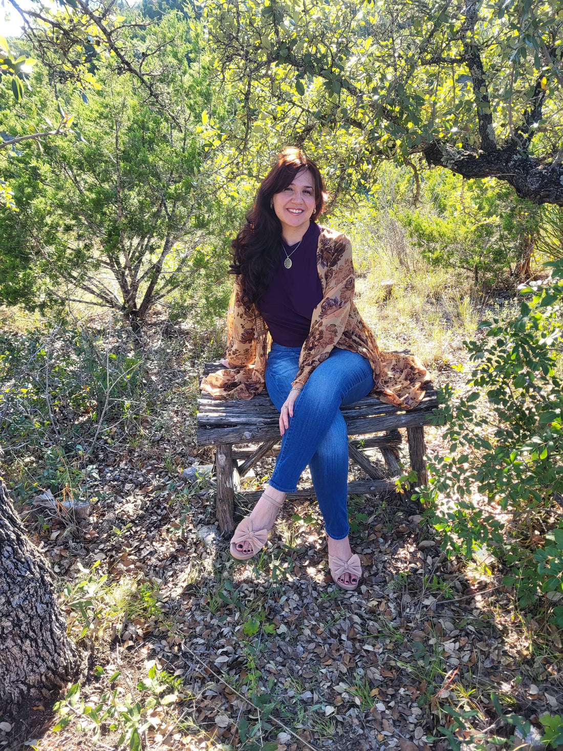 Stephanie Christley, Artist & Founder of SLChristley Designs, sitting outdoors on a wooden bench smiling, surrounded by lush green trees.